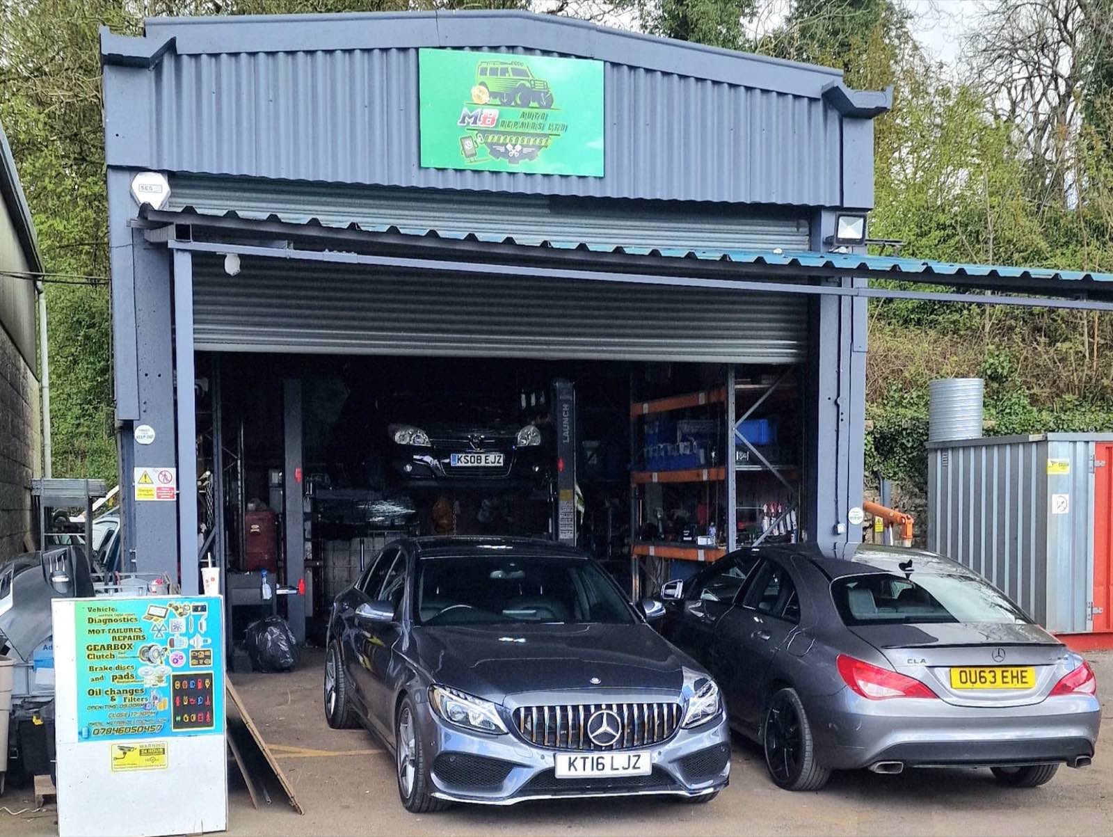 The MB Auto Repairs workshop at Millbrook Depot near Shepton Mallet: a steel-clad unit with the roller shutter up, a car raised on a two-post lift inside, parts racking along the wall, and two Mercedes parked on the forecourt.